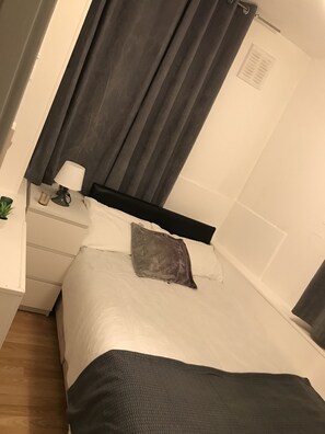 Un lit bien fait, avec une tête de lit noire, une lampe de chevet et une petite plante en pot posée sur une table de chevet blanche.