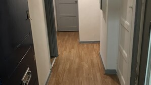 A narrow hallway with wooden flooring, a dark door on the left, and a light-colored door at the end. - Charming 3-bed Flat in London (London)