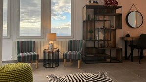 A living room with two striped armchairs, a round side table with a lamp, a bookshelf, and a view of the sky through large windows. - Casa Anna (La Puntilla)
