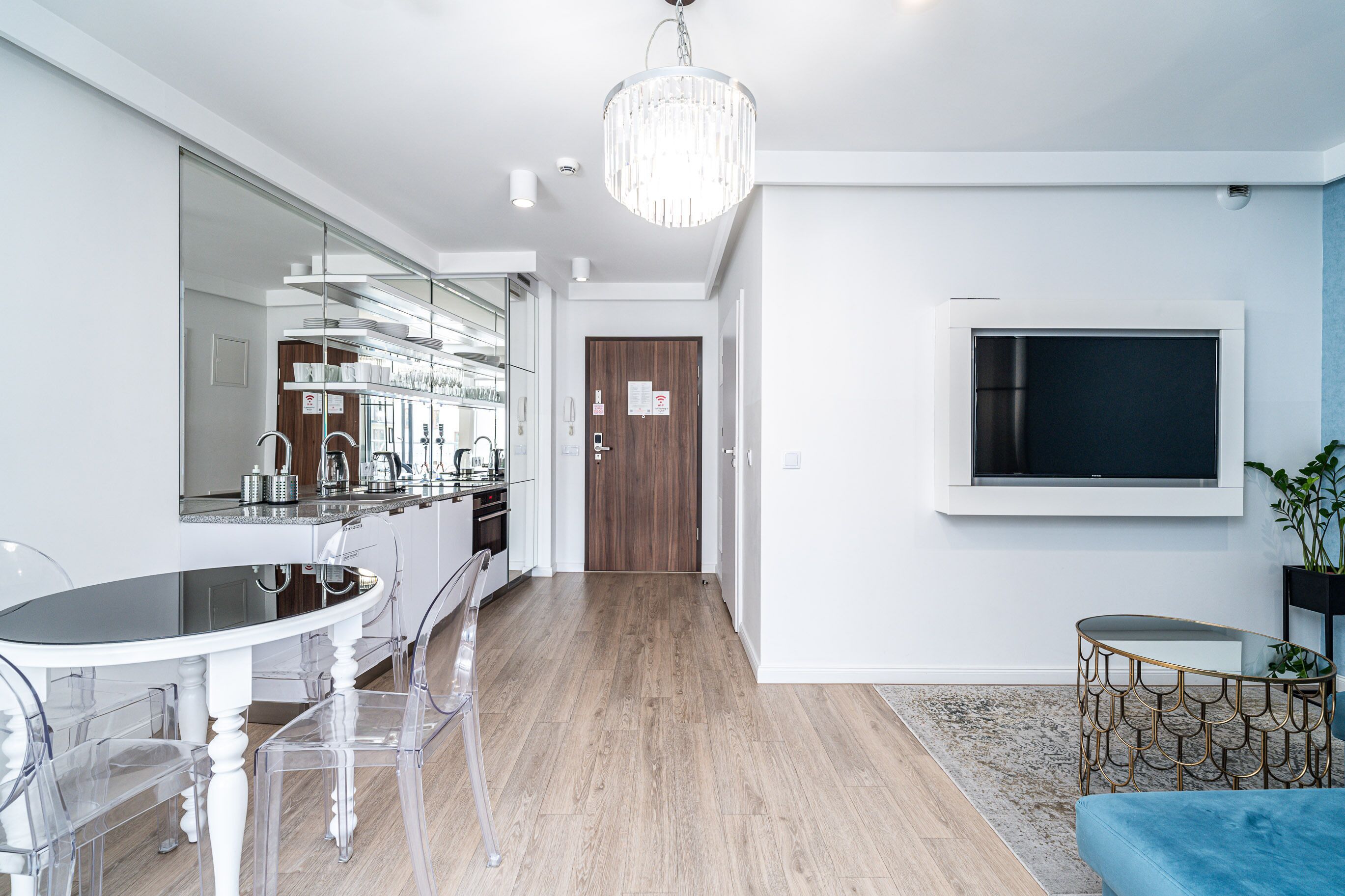 A modern kitchen with a dining area, a TV mounted on the wall, and a hallway leading to a door.