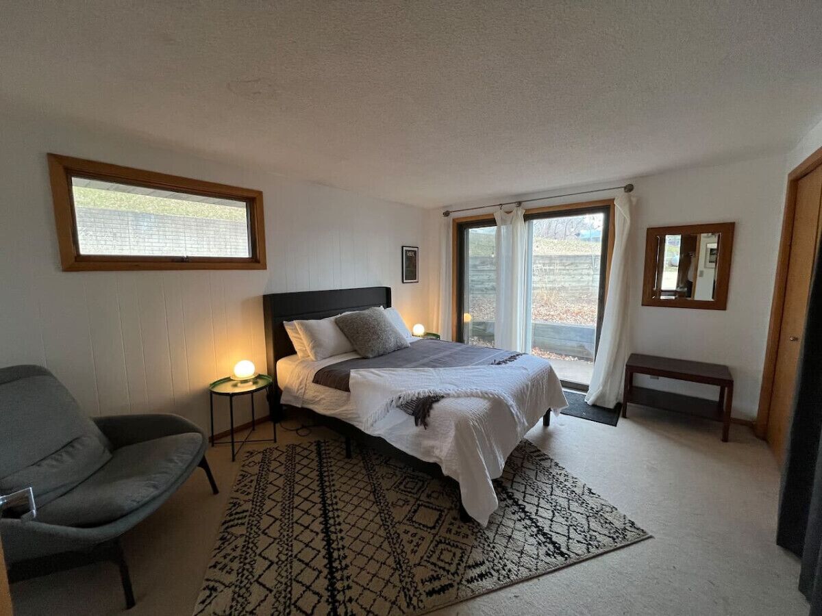 A bedroom with a bed, a chair, a bedside table, a rug, and a window.