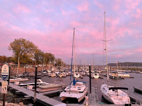 A marina with multiple boats docked at wooden piers, a sign prohibiting rock throwing, and a calm water body in the background.