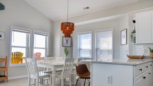 A modern kitchen with white cabinetry, a central island, and a dining area with white chairs and a table. - Sandy By The Sea | Gorgeous Oceanfront Retreat + Private Elevator! (North Topsail Beach)