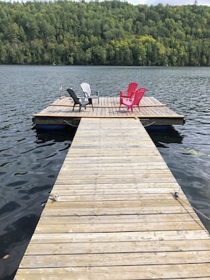 A wooden dock extending into a lake with two Adirondack chairs on one side.