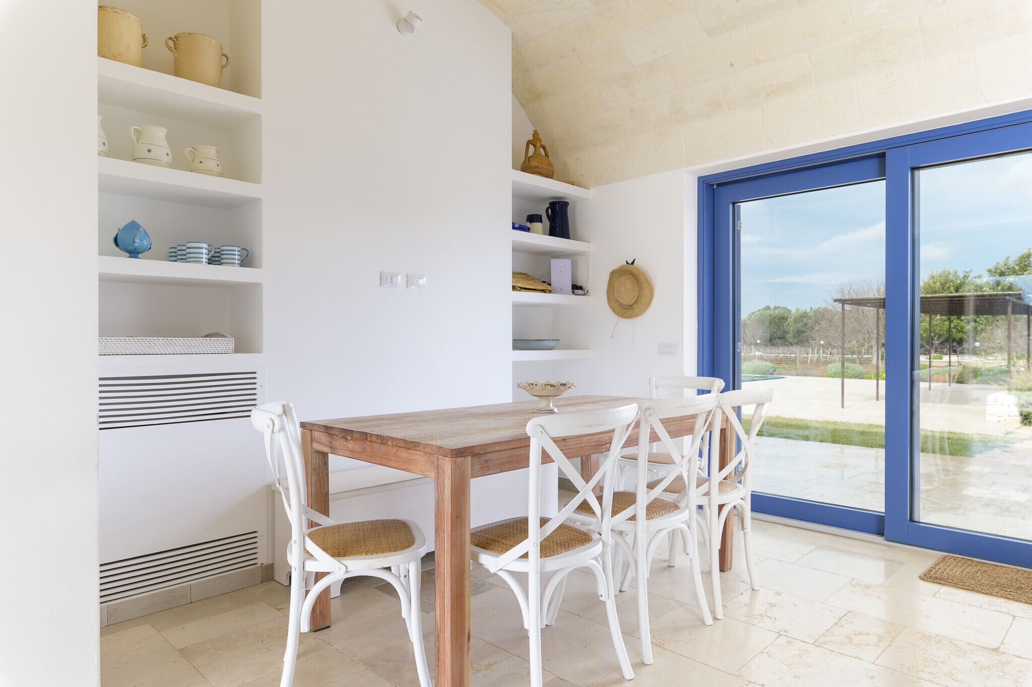 A dining area with a wooden table and white chairs, white shelving with kitchenware, and a large sliding glass door to an outdoor area.