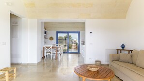 Een ruime woonkamer met een houten tafel en een beige bank, een eethoek met witte stoelen, en een uitzicht door een glazen deur op een terras.