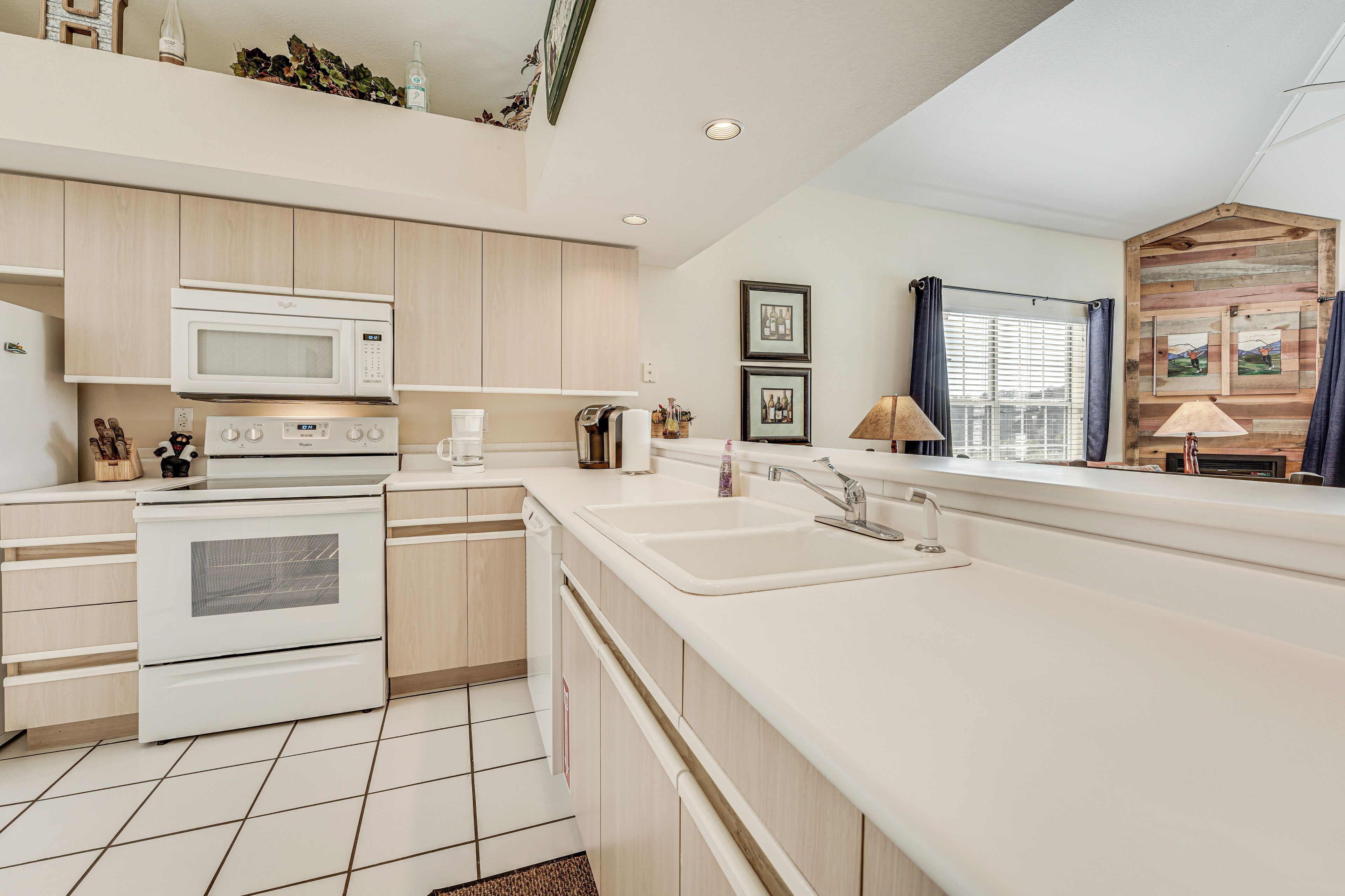 A modern kitchen with white appliances, wooden cabinets, a large sink, and a window with curtains.