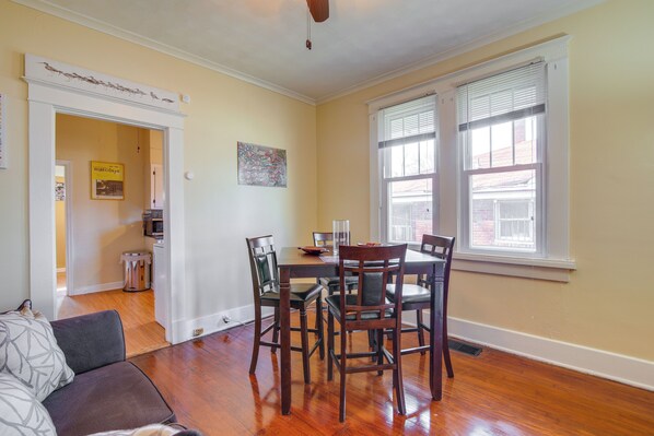 A dining area with a wooden table and chairs, a kitchen in the background, and a window with blinds.