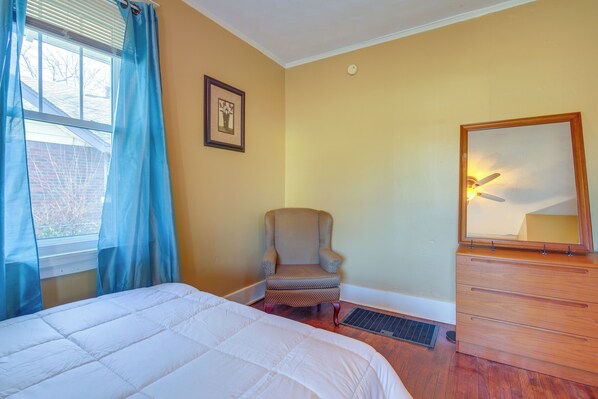 Un dormitorio con cama, una silla, un aparador y una ventana con cortinas.