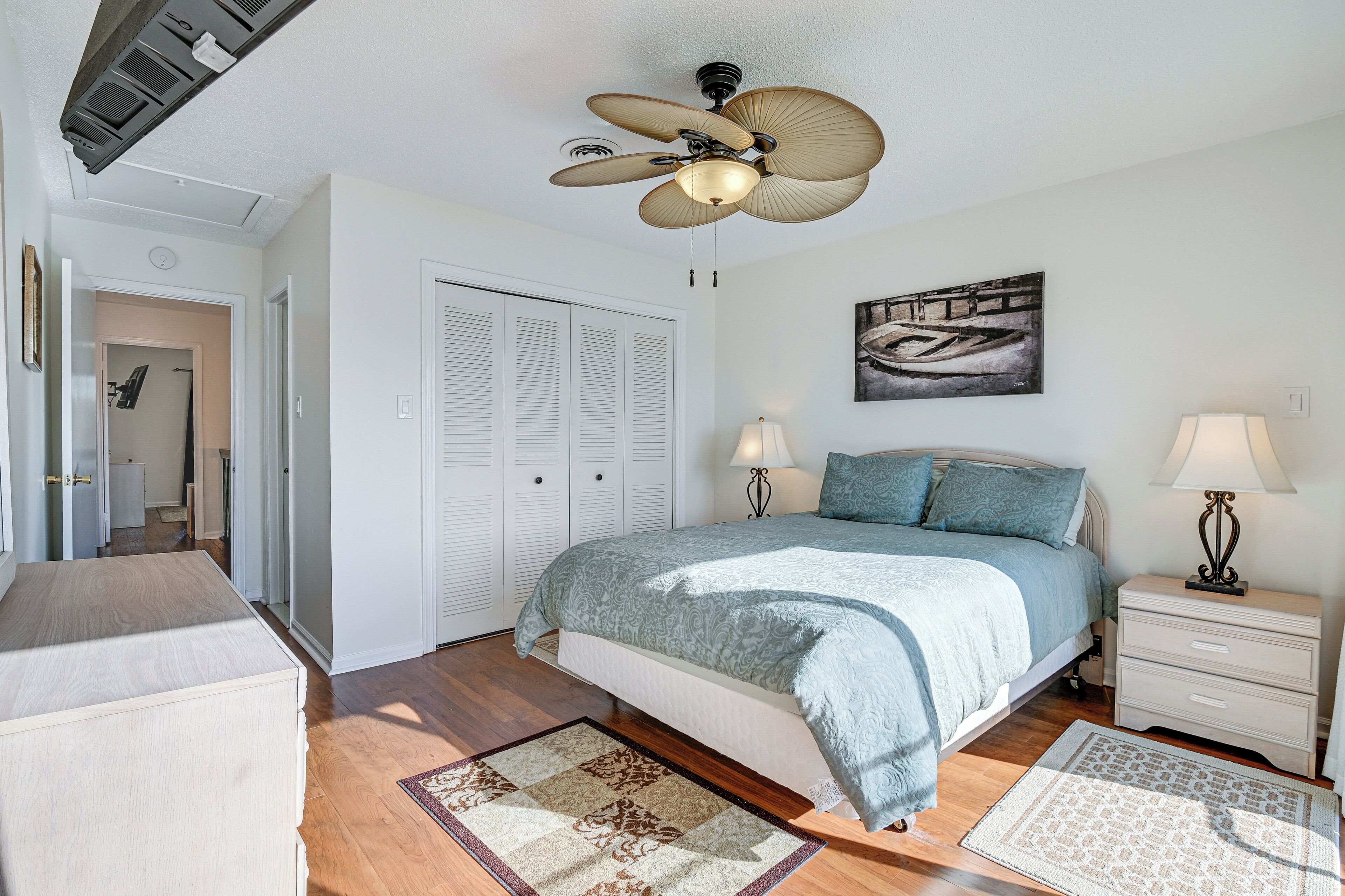 Une chambre à coucher comprenant un lit, deux tables de chevet avec des lampes, un ventilateur de plafond, une porte et une œuvre d’art accrochée au mur.