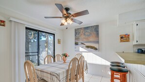 Un coin repas avec une table dressée pour quatre personnes, un ventilateur de plafond et un espace cuisine comprenant un comptoir et des tabourets.