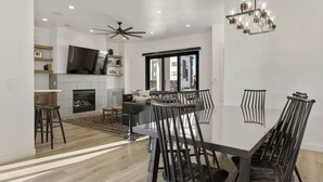 A modern dining area with a glass-top table, black chairs, a chandelier, a fireplace, and a ceiling fan.