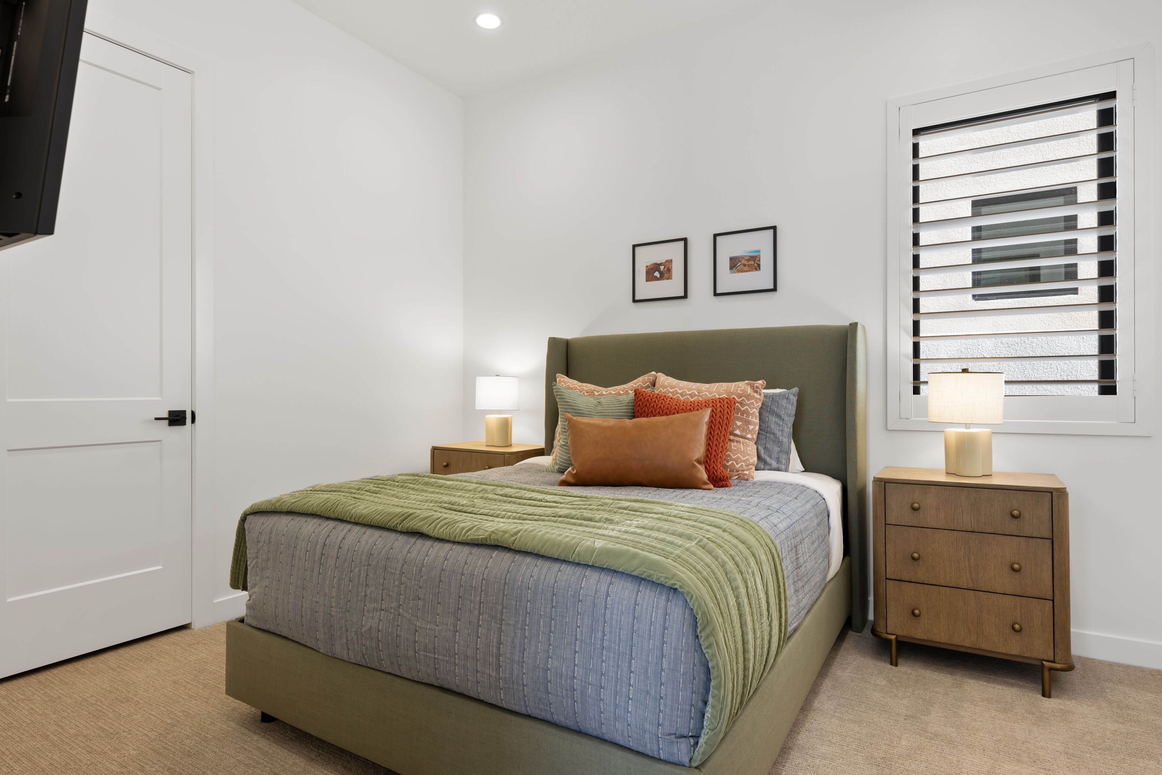 Ein Schlafzimmer mit Bett, Nachttischen, Kommode, Fenster mit Jalousien und zwei gerahmten Bildern an der Wand.