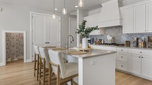 A modern kitchen with white cabinetry, a central island with bar stools, pendant lighting, and a backsplash with a geometric pattern. - Mermaid Lagoon 4 Bedroom Townhouse (St. George)