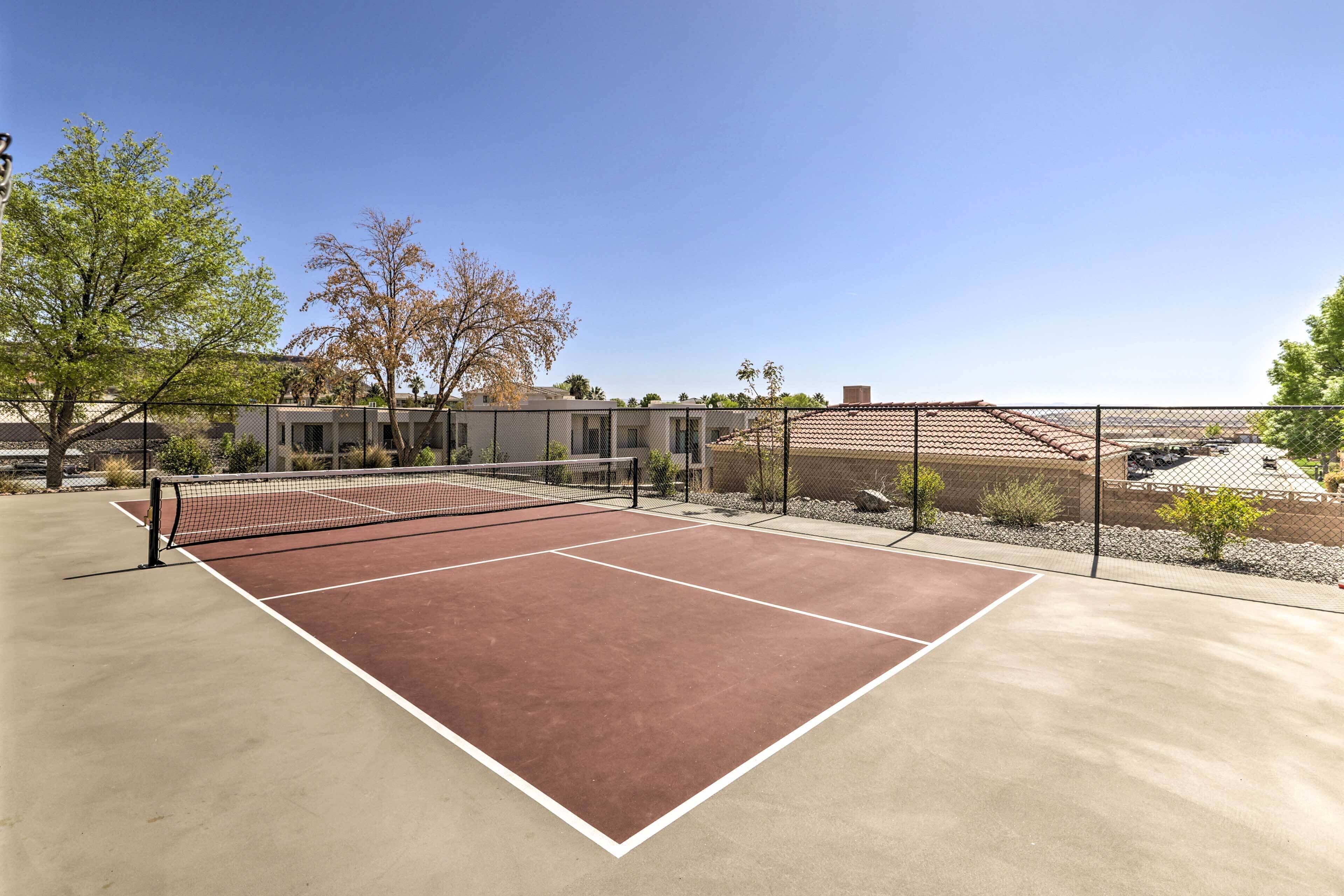 An outdoor tennis court with a net, surrounded by a fence and trees.