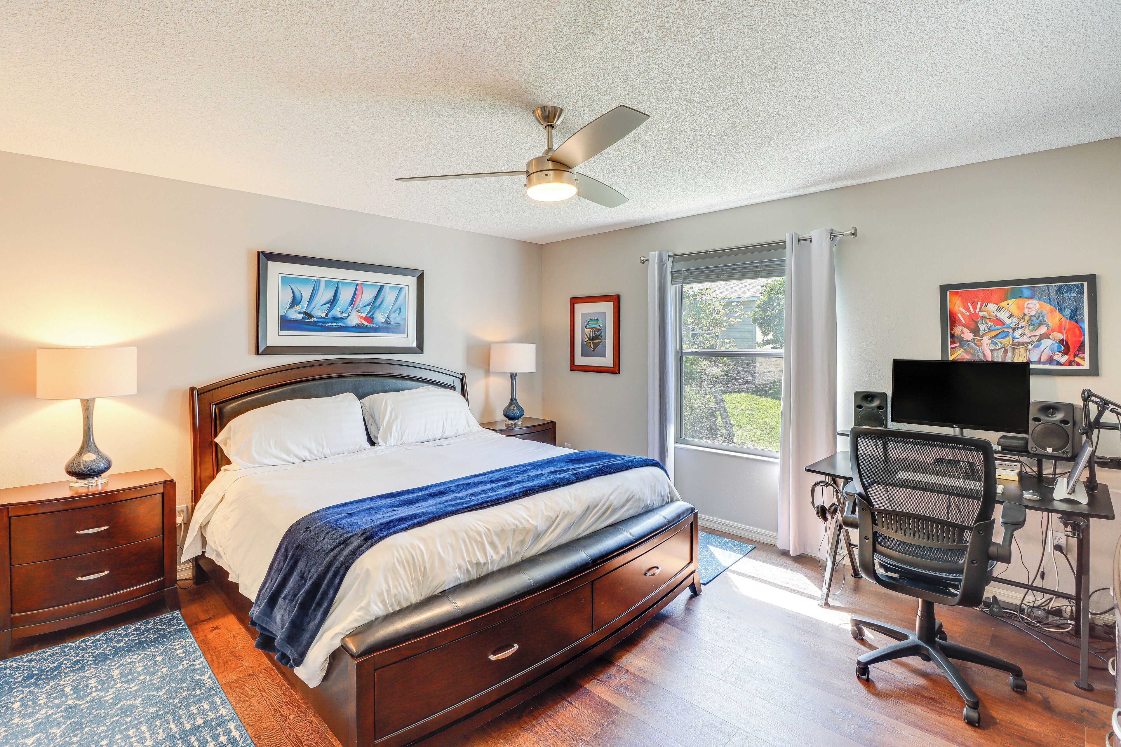 Quarto com cama, escrivaninha com computador, ventilador de teto e obras de arte nas paredes.
