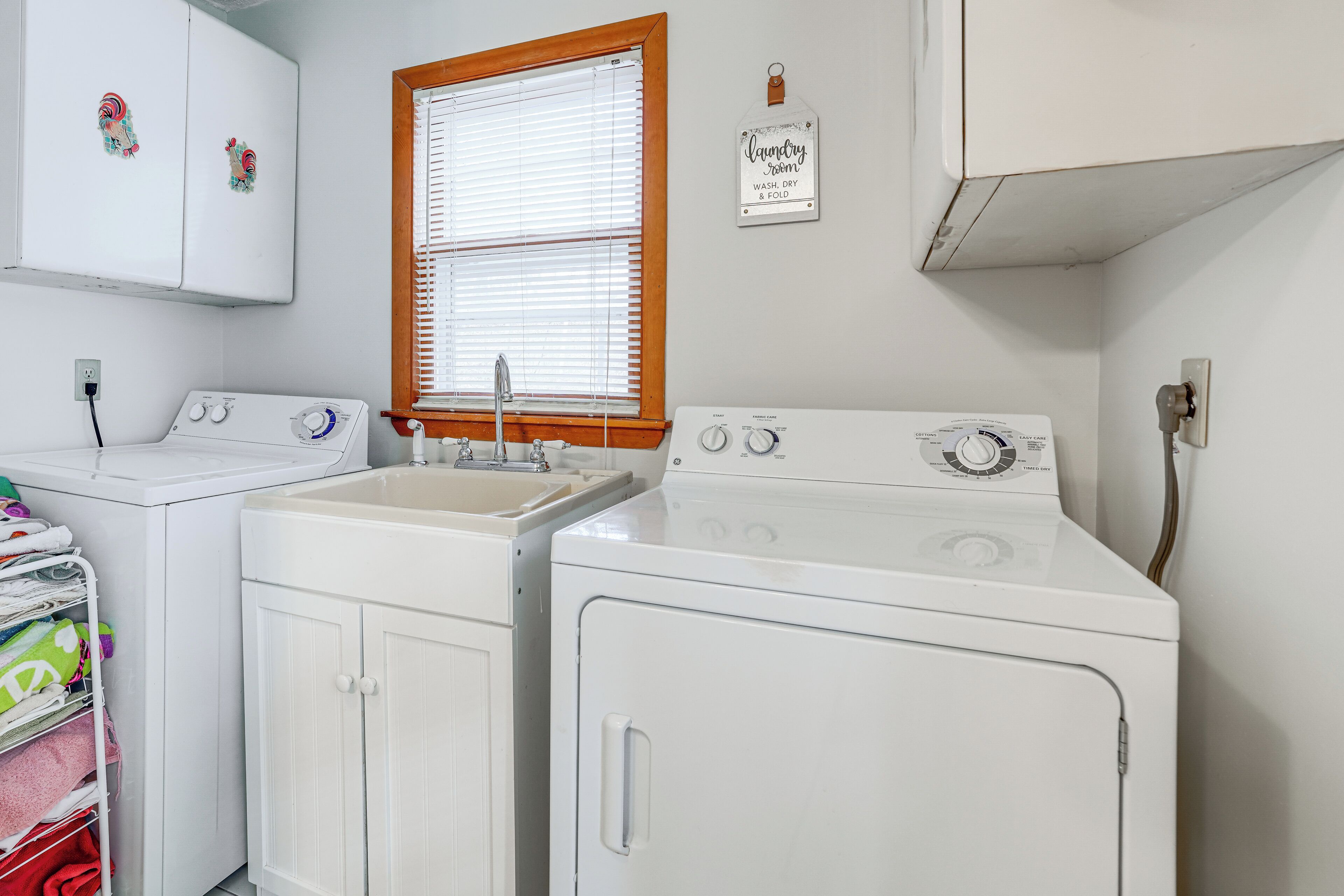 A laundry room with two front-loading washing machines, a sink, and a window with blinds.