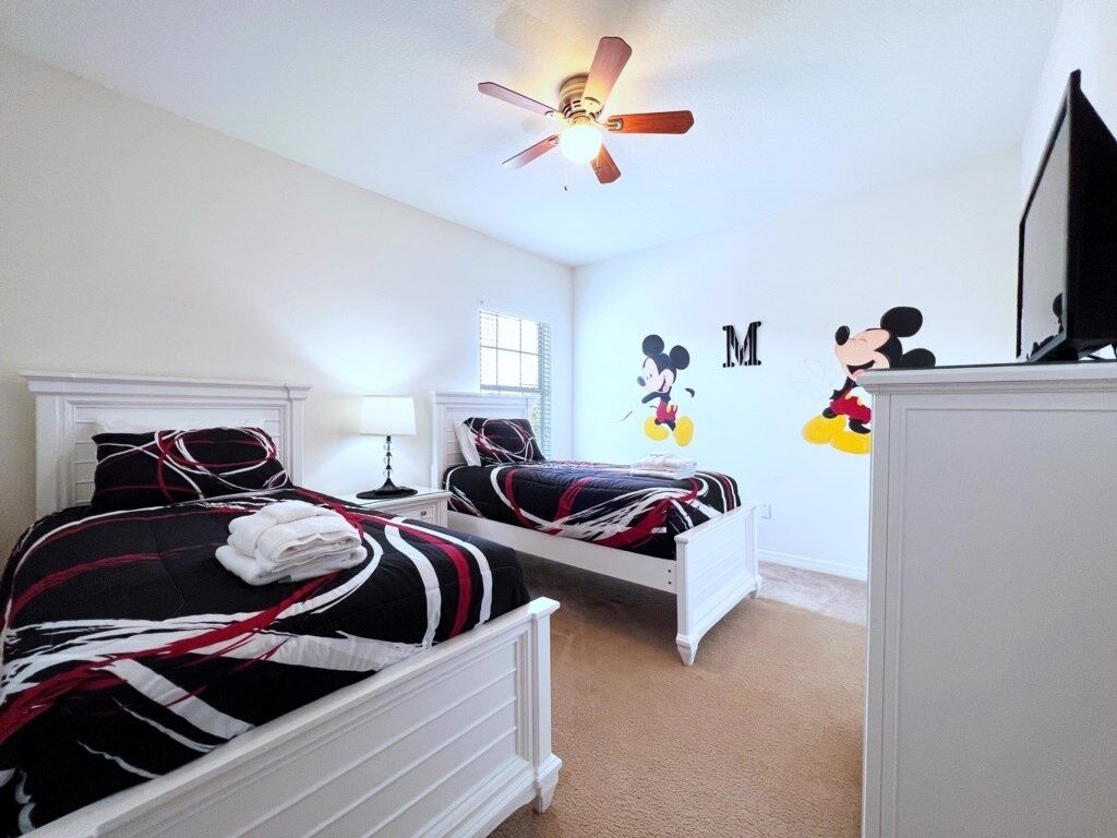 A bedroom with two beds, a ceiling fan, a TV, and Mickey Mouse decorations.