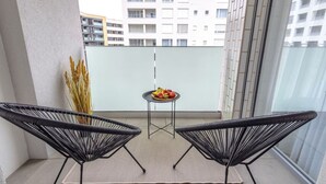 Uma varanda com duas cadeiras de arame preto, uma pequena mesa redonda com frutas e vista para um edifício ao fundo.