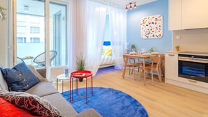 Um apartamento moderno com sala de estar, área de jantar e cozinha compacta. Possui uma janela grande com cortinas leves, um tapete azul e uma pintura abstrata colorida na parede.
