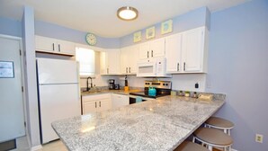 A modern kitchen with white cabinets, a granite countertop, and a built-in oven. There are two bar stools and a coffee maker on the counter. - Seabay Vista II 102 2 Bedroom Condo by RedAwning (Ocean City)