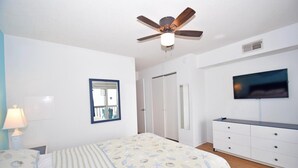 A bedroom with a bed, a ceiling fan, a TV, a dresser, and a window with a view of the outside. - Seabay Vista II 102 2 Bedroom Condo by RedAwning (Ocean City)
