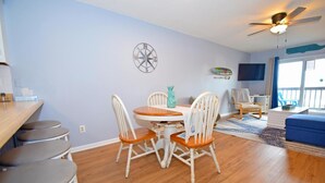 A dining area with a round table and four chairs, a ceiling fan, and a wall-mounted compass. - Seabay Vista II 102 2 Bedroom Condo by RedAwning (Ocean City)