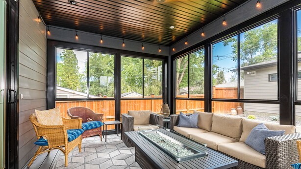 Katettu terassialue, jossa on rottinkihuonekaluja, lasitasoinen sohvapöytä ja suuret ikkunat, joista avautuu näkymä takapihalle.