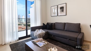 A modern living room with a grey sofa, a coffee table with a book and decorative items, a large window with curtains, and two framed artworks on the wall.