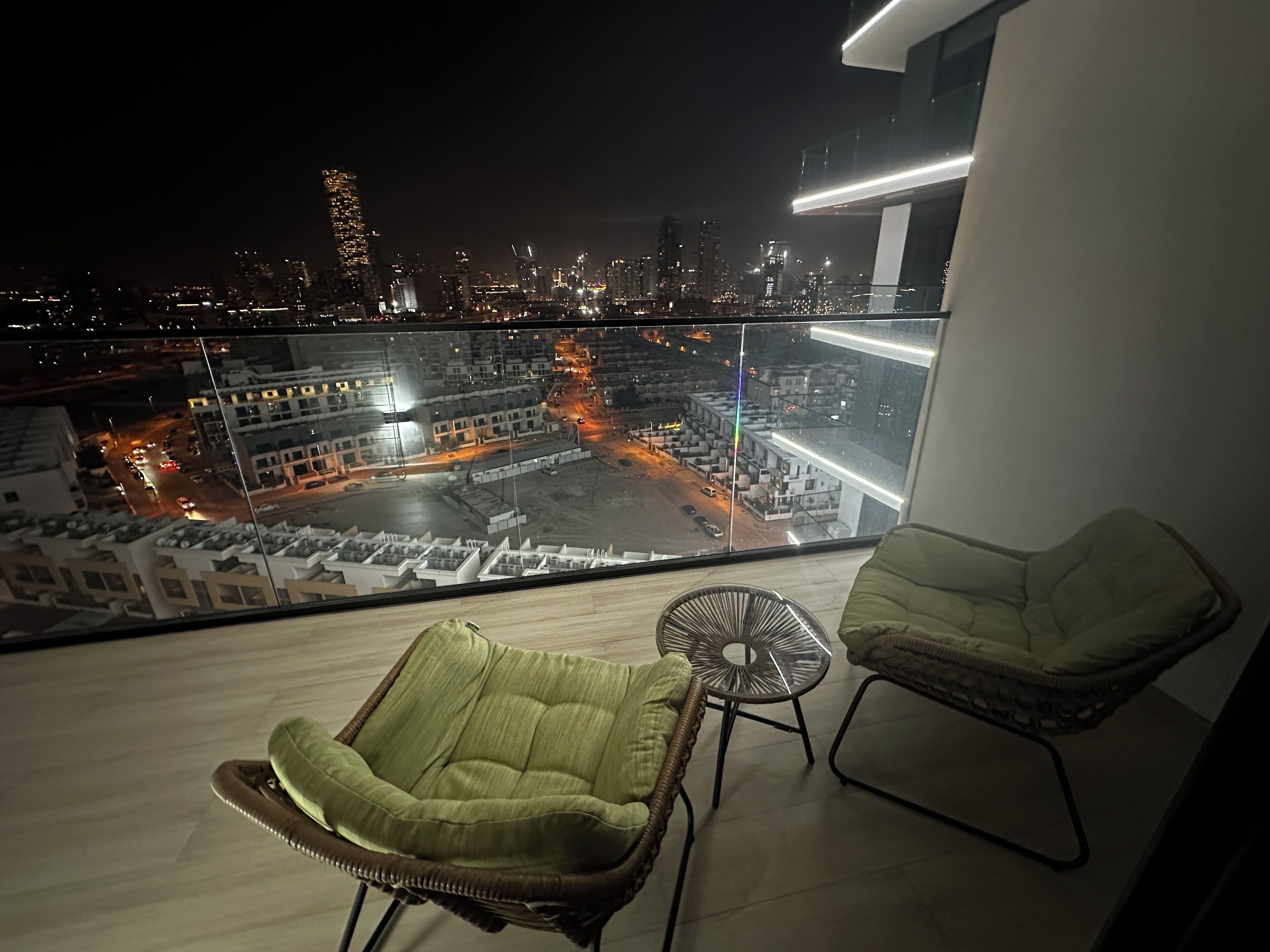 A balcony with two green cushioned chairs and a small table, overlooking a cityscape at night.