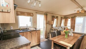 A compact kitchen with a sink, a dining area with a table and chairs, and a view of the outdoors through a window. - A La Mer (Paignton)