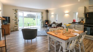 A modern kitchen with white cabinets, a dining area with a wooden table and white chairs, and a cozy living room with a sofa and fireplace. - The Barn (Bishop Auckland)