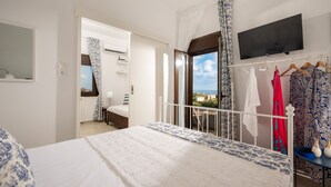 Un dormitorio con cama, televisor montado en la pared, balcón con vistas y un perchero con varias prendas.