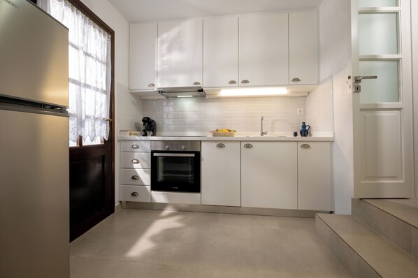 A modern kitchen with white cabinets, a stainless steel refrigerator, a built-in oven, and a window with lace curtains. - Marianthi's Home is ensconced in the enchanting Village of Old Hersonissos. (Chersonisos)
