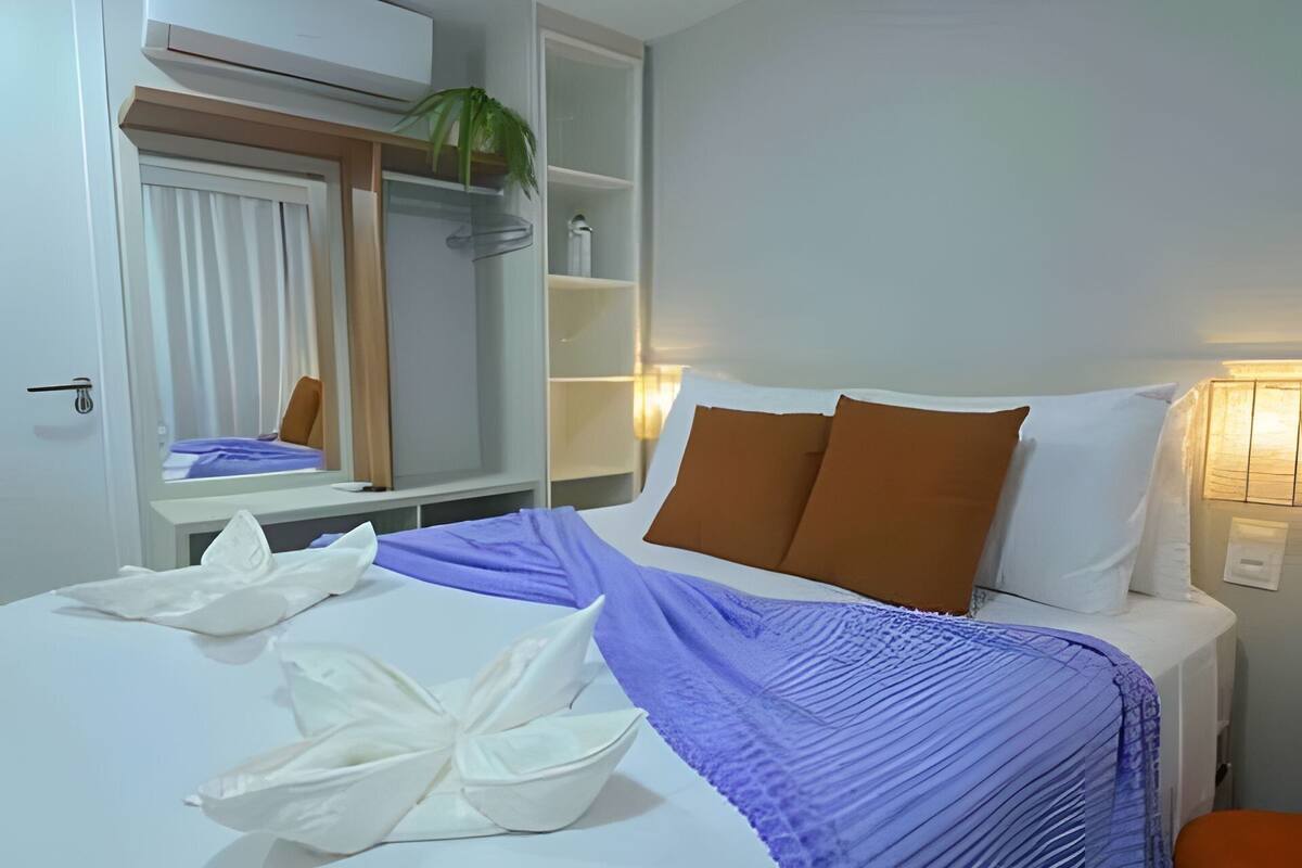 Una habitación de hotel con cama, escritorio, espejo, repisa con artículos de aseo y una planta en maceta.