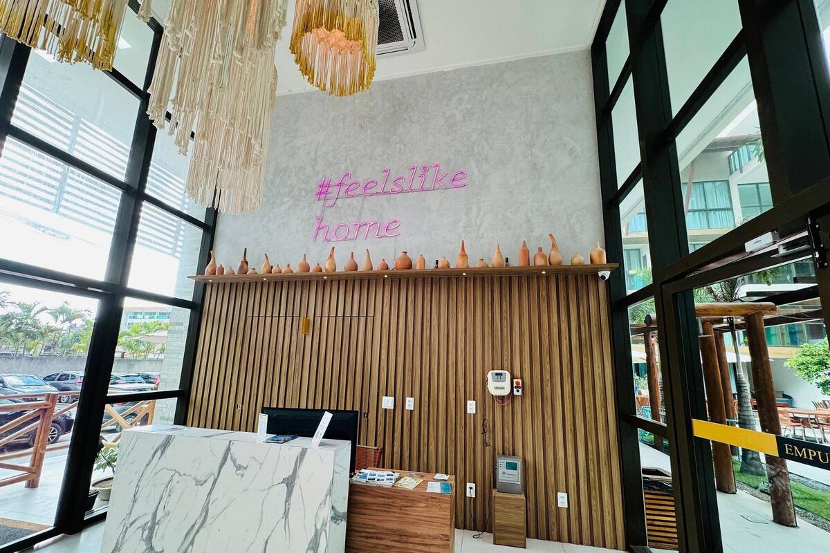 Un mostrador de recepción con encimera de mármol, una pared de madera con listones verticales y un letrero de neón que dice '#feelslike home'.