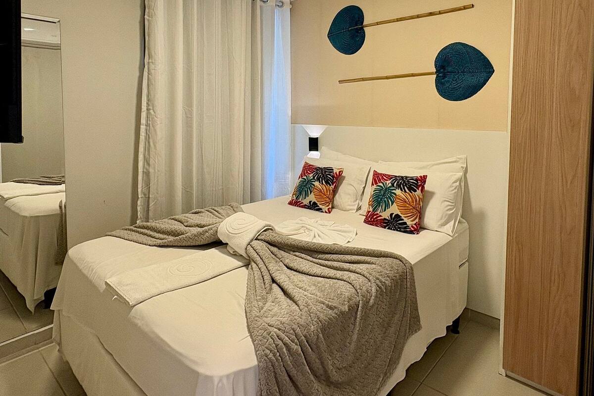 Una cama bien hecha con una colcha, una mesita de noche, un espejo y una pared con dos adornos decorativos.
