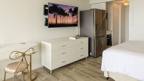 A hotel room with a bed, a white dresser, a desk with a chair, a refrigerator, and a painting of palm trees on the wall. - Glam Condo Located in Central Waikiki, Panoramic Ocean View, & Full Kitchen!
 (Honolulu)