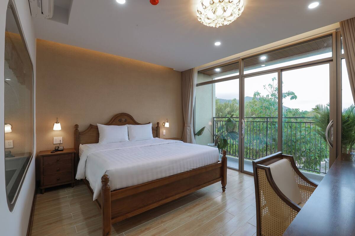 A bedroom with a large bed, bedside tables, a chair, and a balcony with a view of greenery.