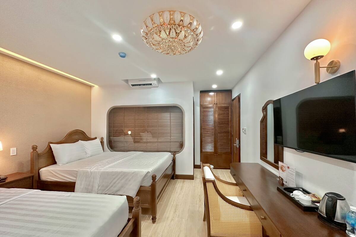 A hotel room with two beds, a TV, a desk with a kettle, and a chandelier.