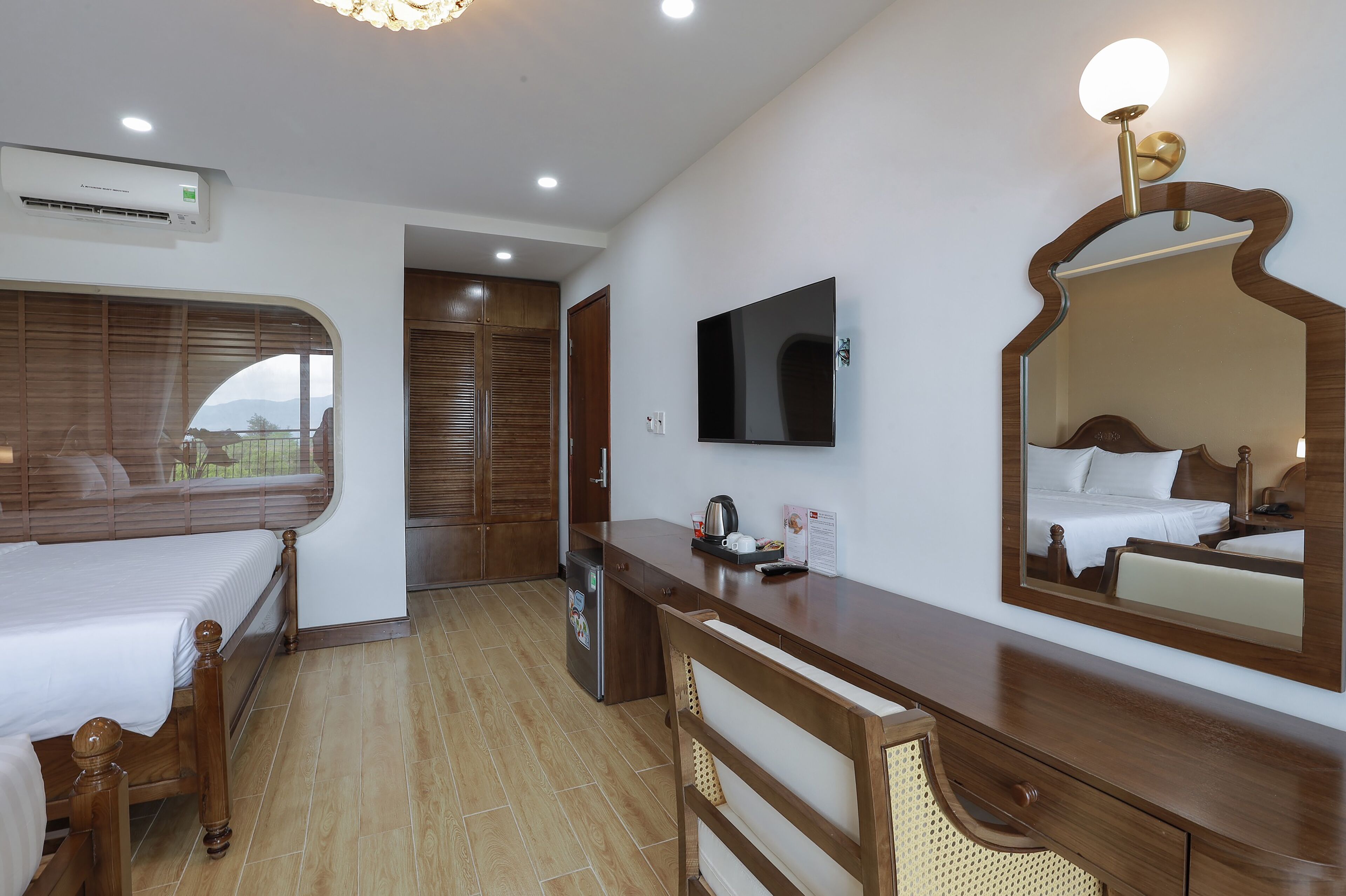 A hotel room with a bed, a desk, a mirror, a TV, and an air conditioning unit.