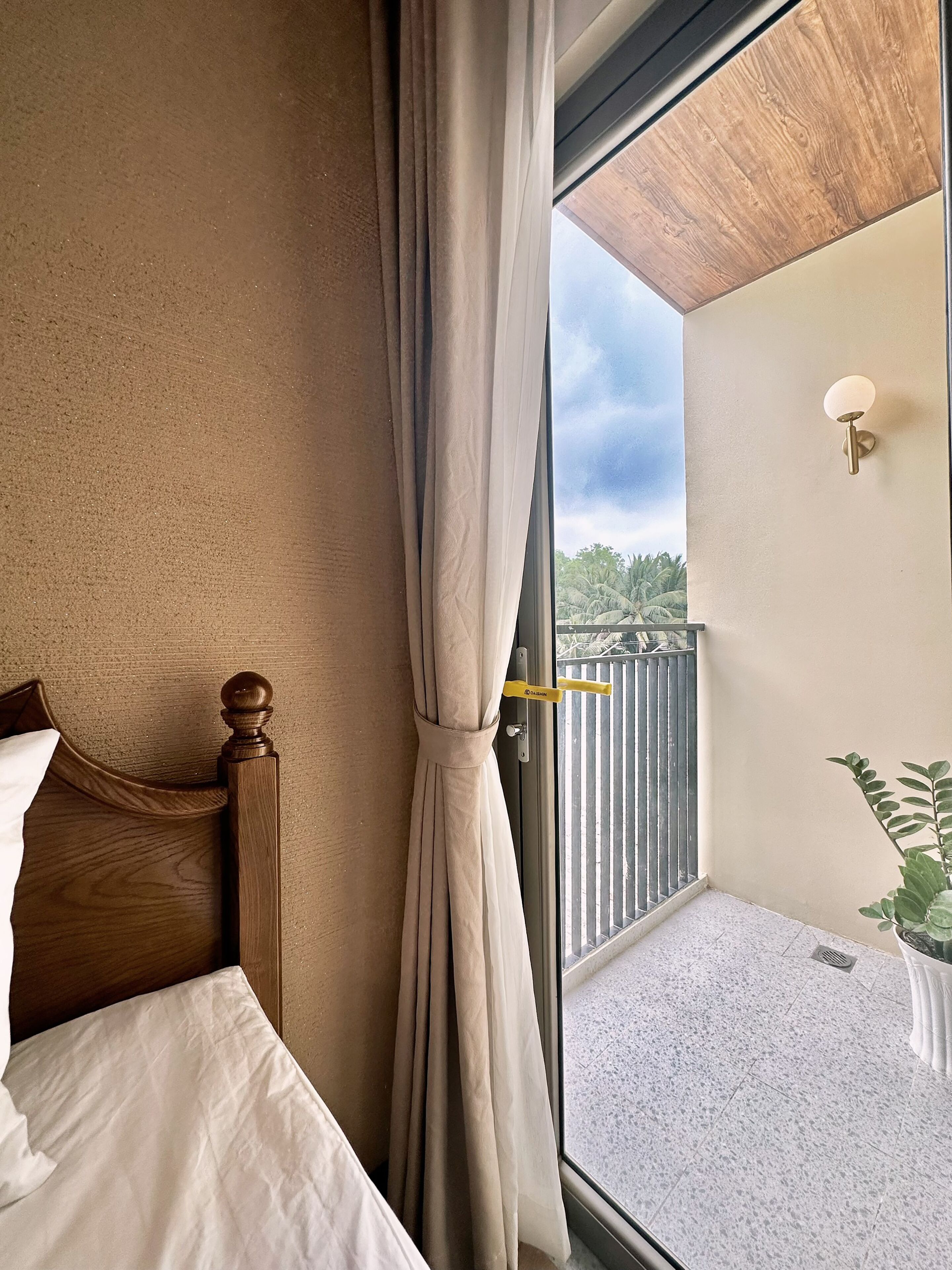 Un dormitorio con cama, cortina, balcón con barandilla y planta en maceta.