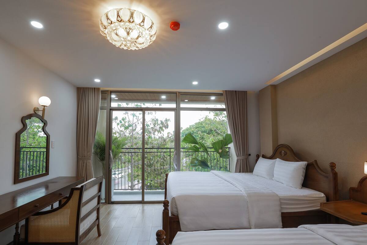 A hotel room with two beds, a desk, a chair, a chandelier, and a balcony with a view of greenery.