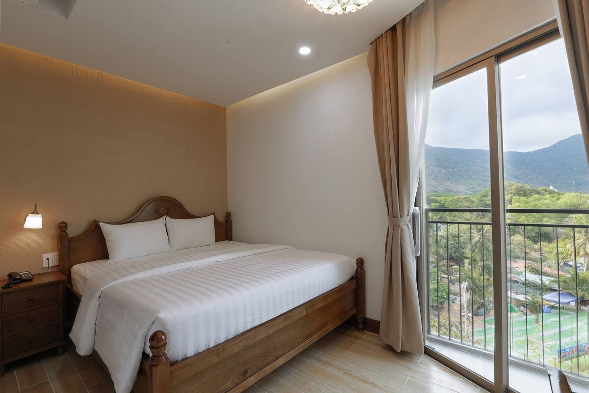 A bedroom with a wooden bed, a nightstand, a bedside lamp, and a large window with a view of greenery and mountains.