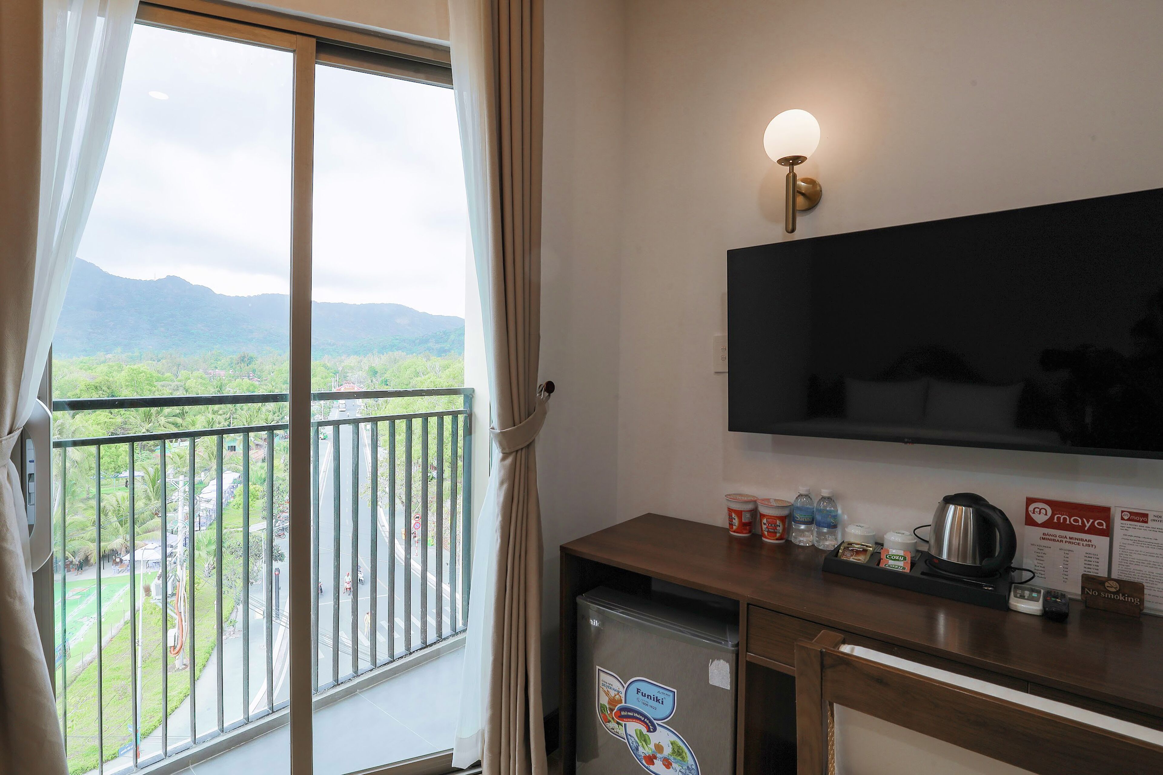 Habitación de hotel con balcón, minibar, televisión y vistas a un paisaje verde.