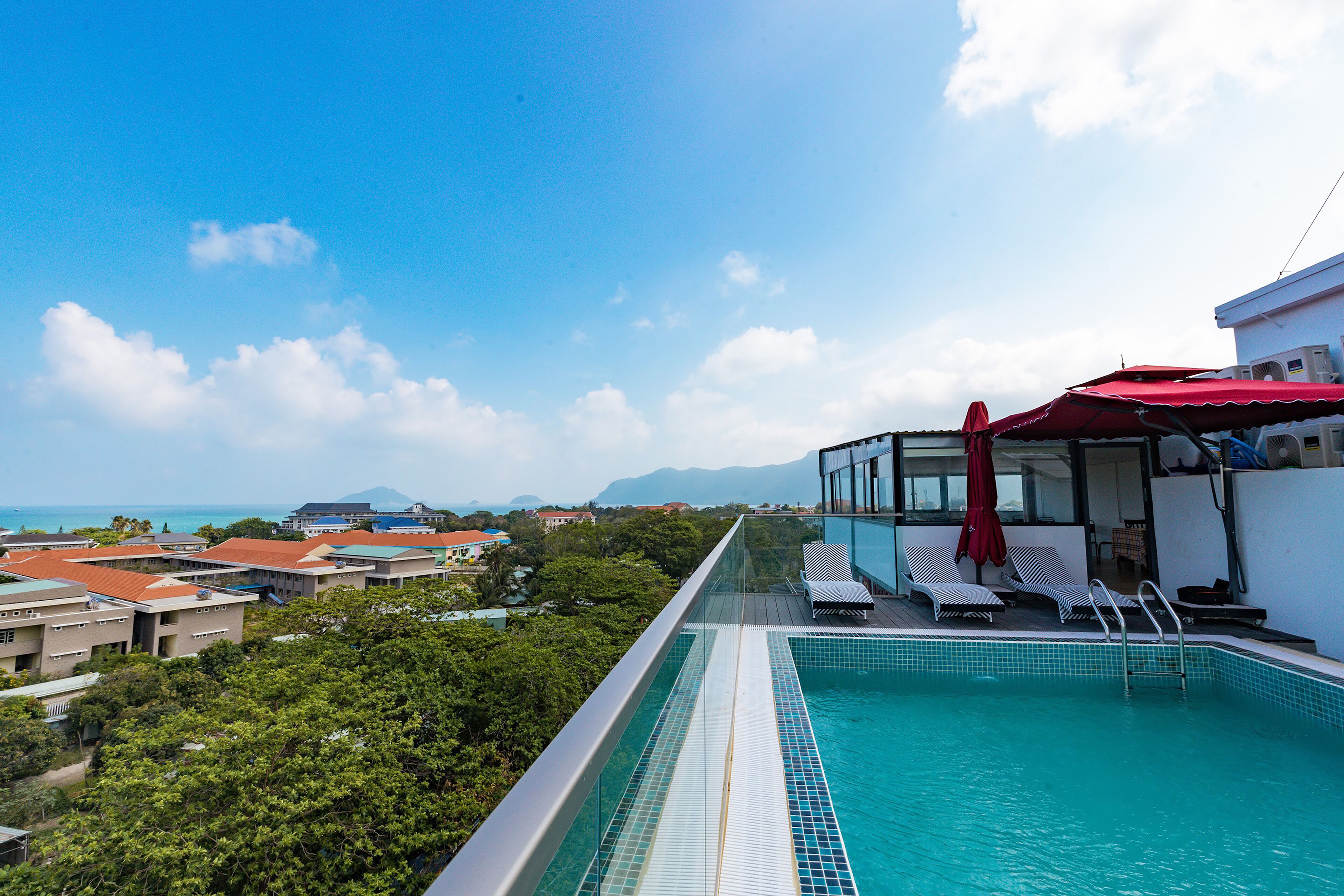 Piscina infinita en la azotea con vistas al océano y montañas, sillas de descanso y una sombrilla roja.