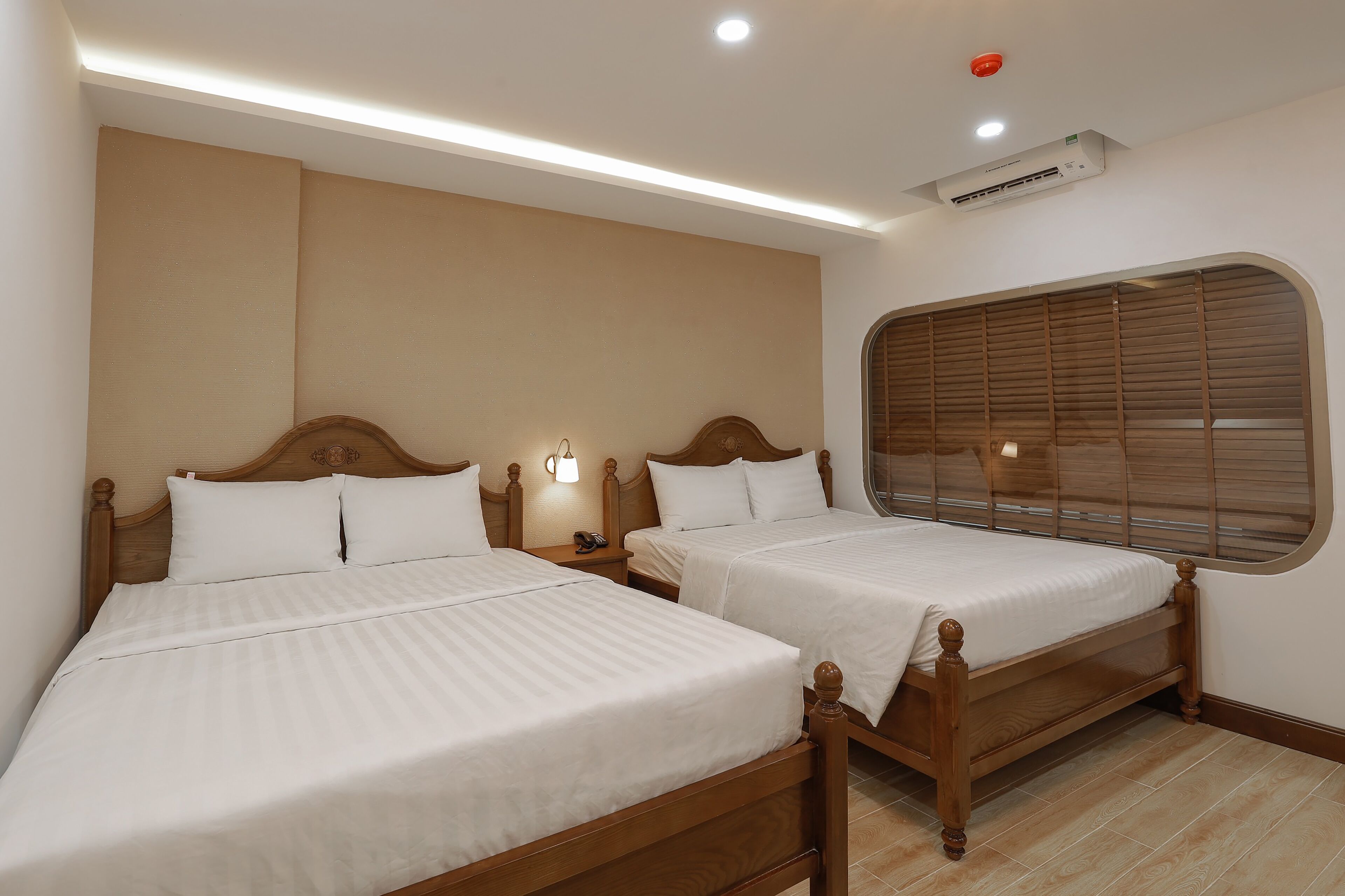 A hotel room with two wooden beds, a nightstand with a lamp, a window with blinds, and an air conditioning unit.