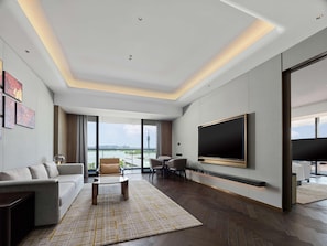 A modern living room with a large flat-screen TV, a sofa, a coffee table, and a view of a cityscape through large windows. - Hilton Changshu (Suzhou)