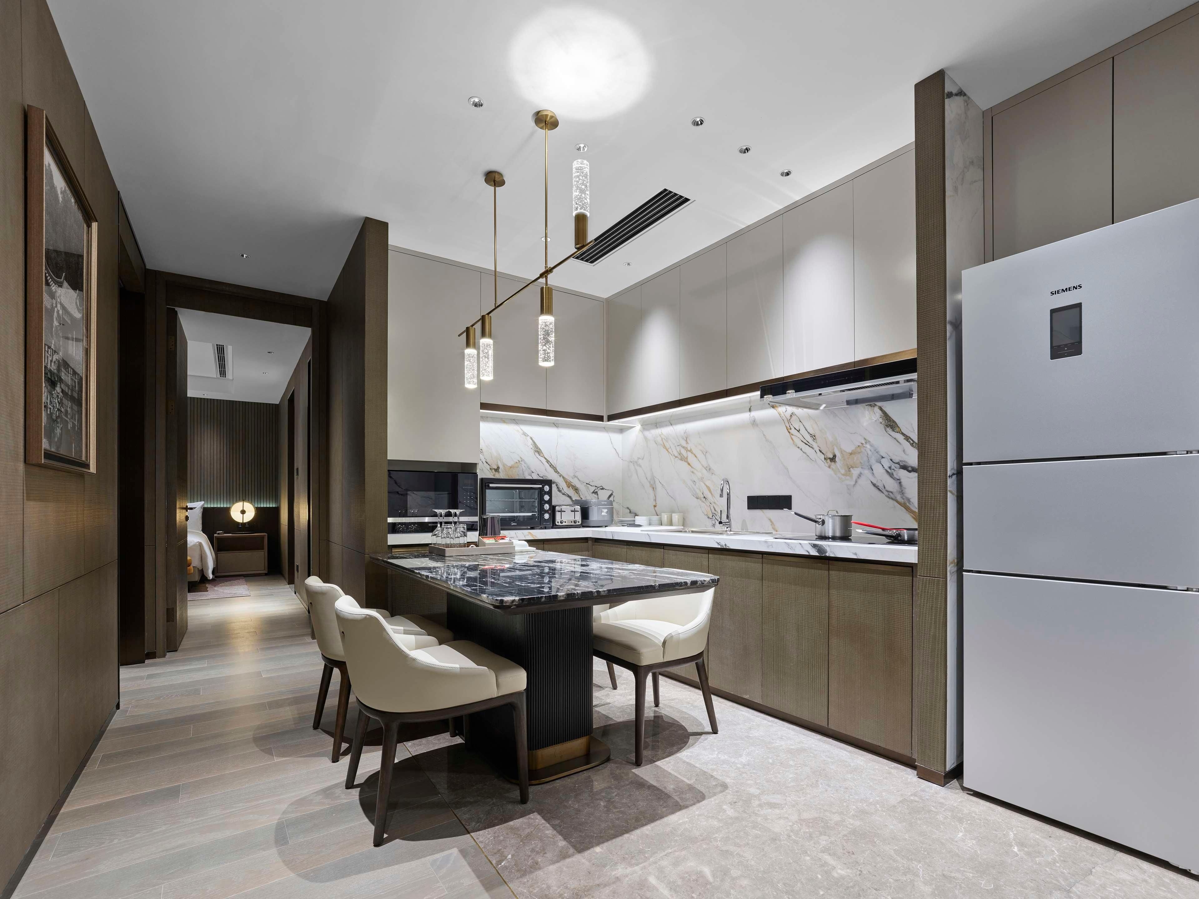 Una cocina moderna con encimera de mármol, dos sillas blancas, un refrigerador empotrado e iluminación colgante.