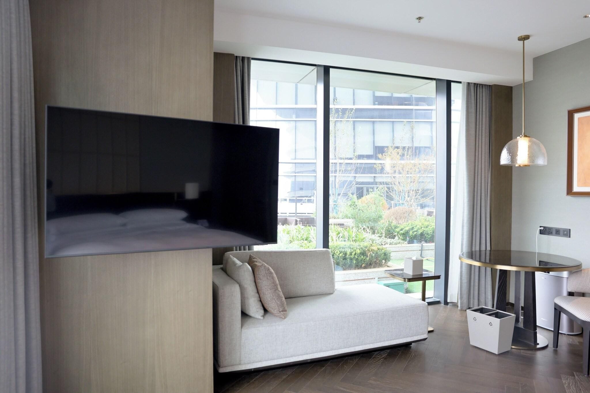 Una habitación de hotel moderna con un ventanal grande, un televisor de pantalla plana en la pared, un sofá, una mesita pequeña y una lámpara colgante.